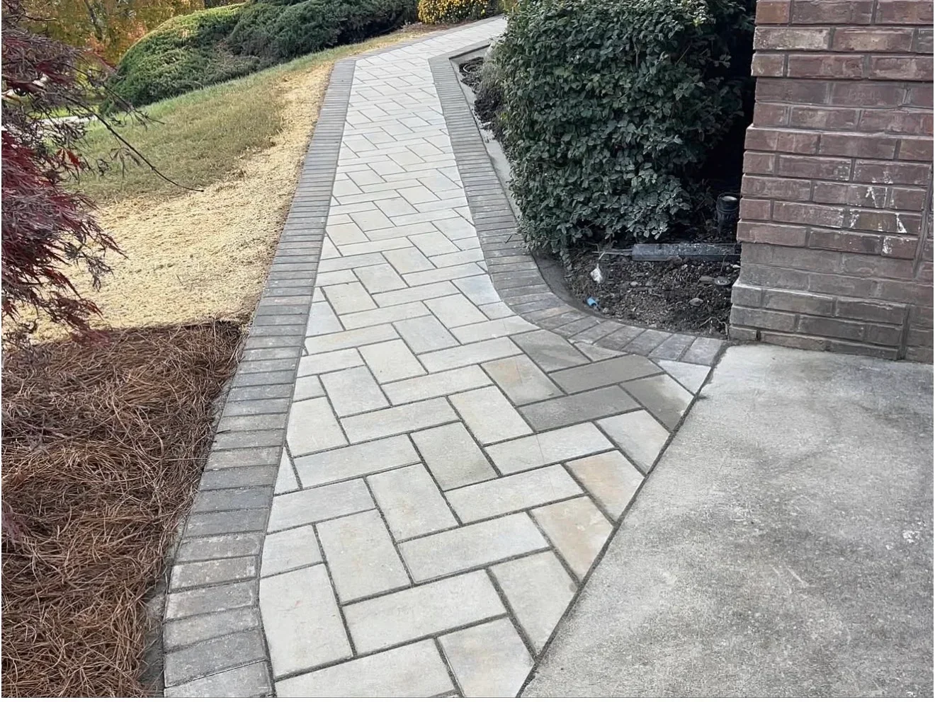 Curved walkway of light gray rectangular pavers with dark gray paver border edging beside a brick home and concrete driveway