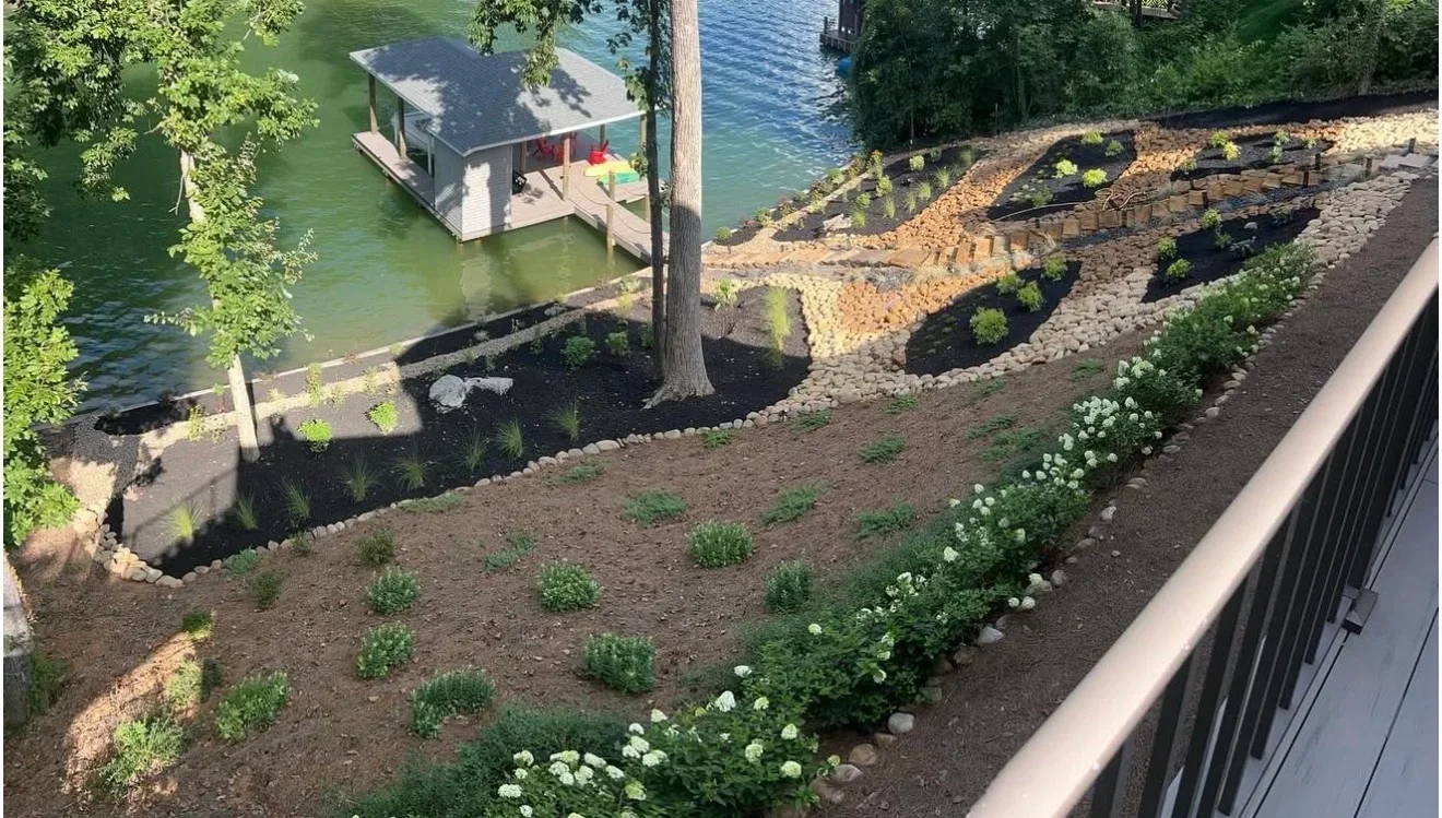 Sloped lakefront landscape with dark mulch beds, white hydrangeas, river rock borders, and timber-edged terraces