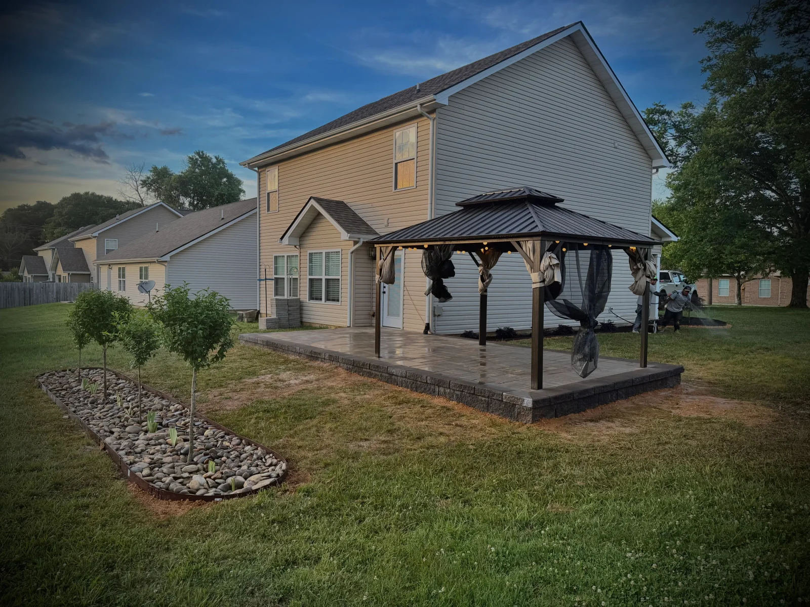 Paver patio with metal-roof gazebo and curved river rock bed with young trees beside vinyl-sided home
