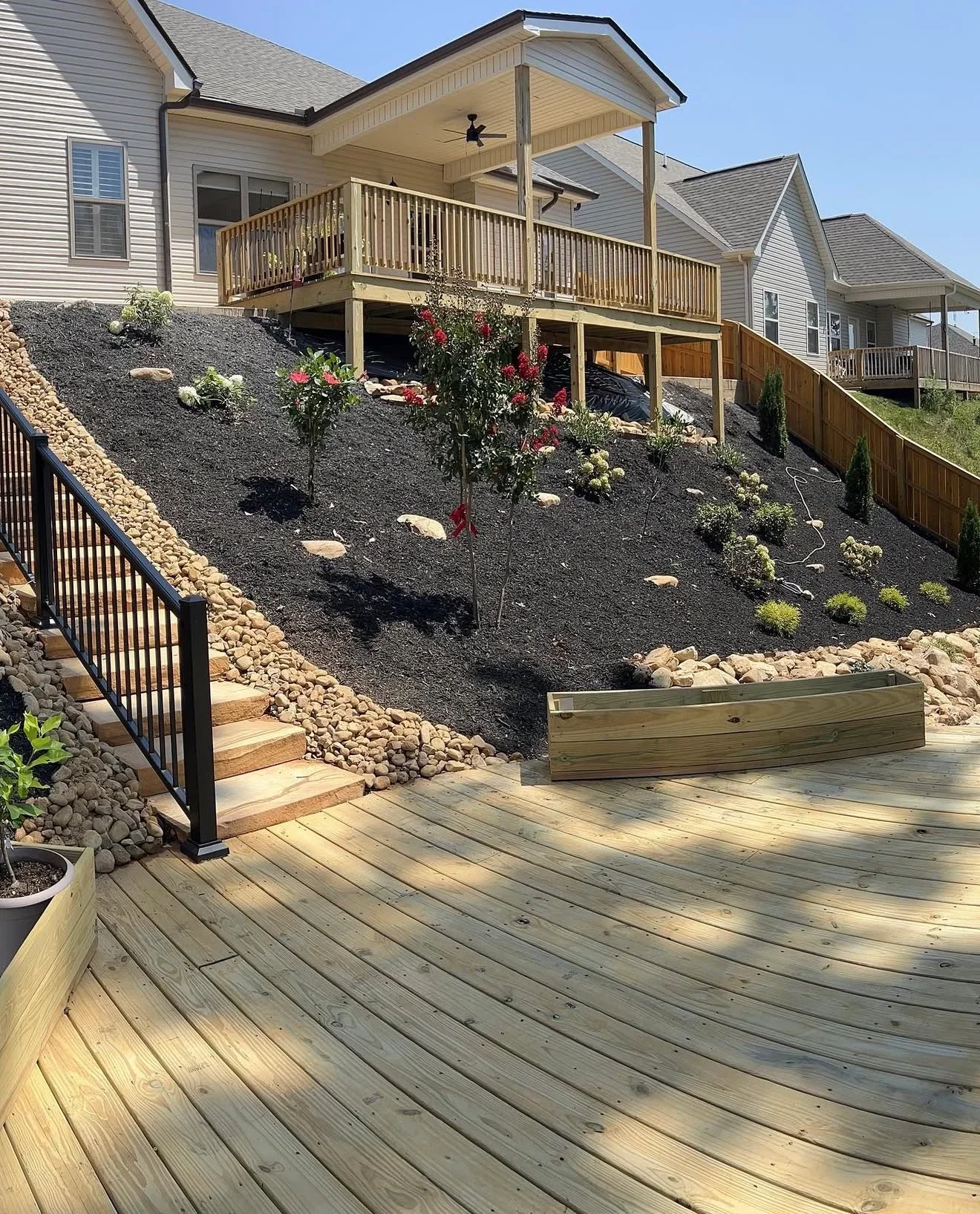 Sloped backyard with dark mulch, river rock, stone steps, black metal railing, and pressure-treated wood deck and planter