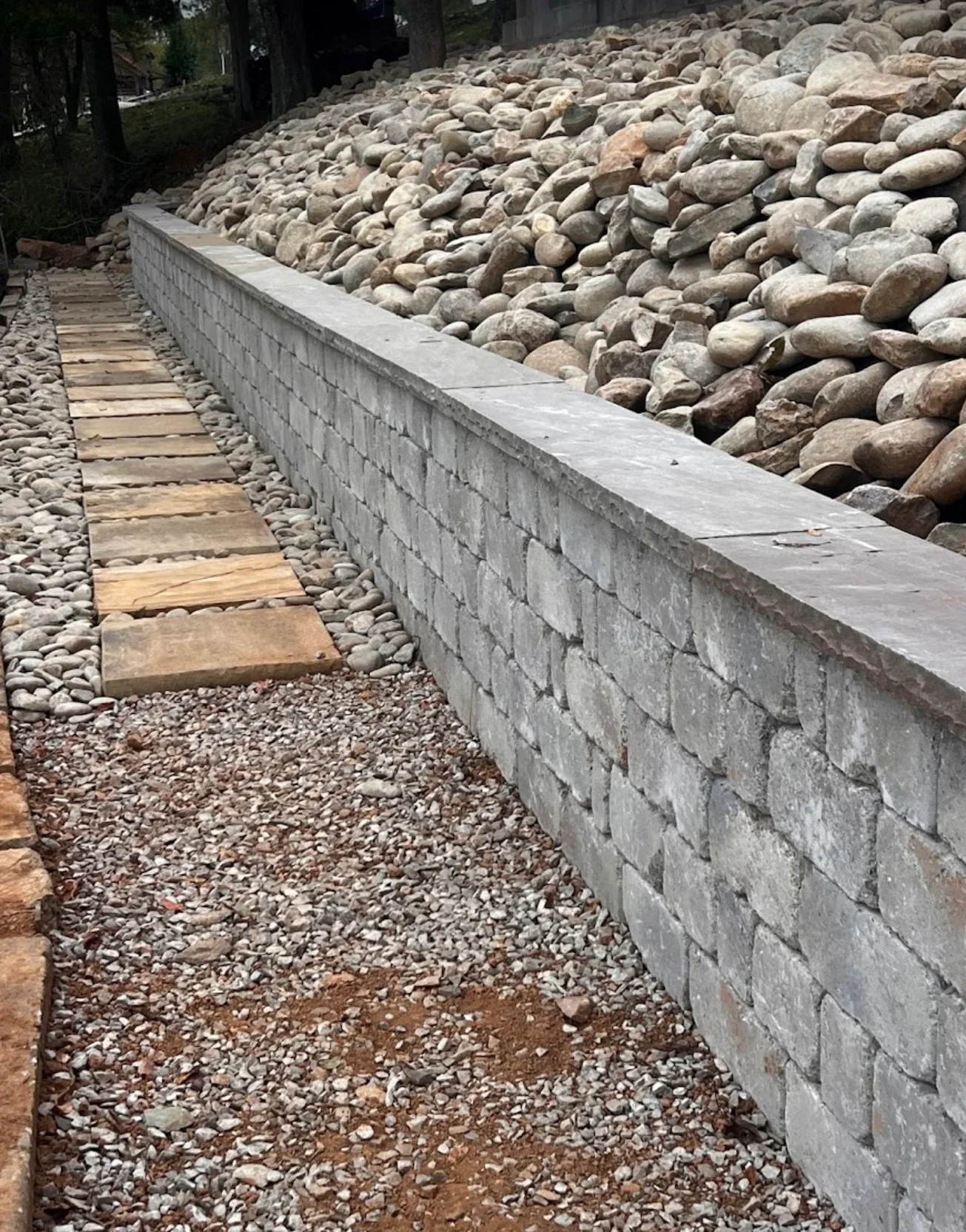 Gray stacked block retaining wall with cap stones, river rock backfill, and flagstone walkway over pea gravel