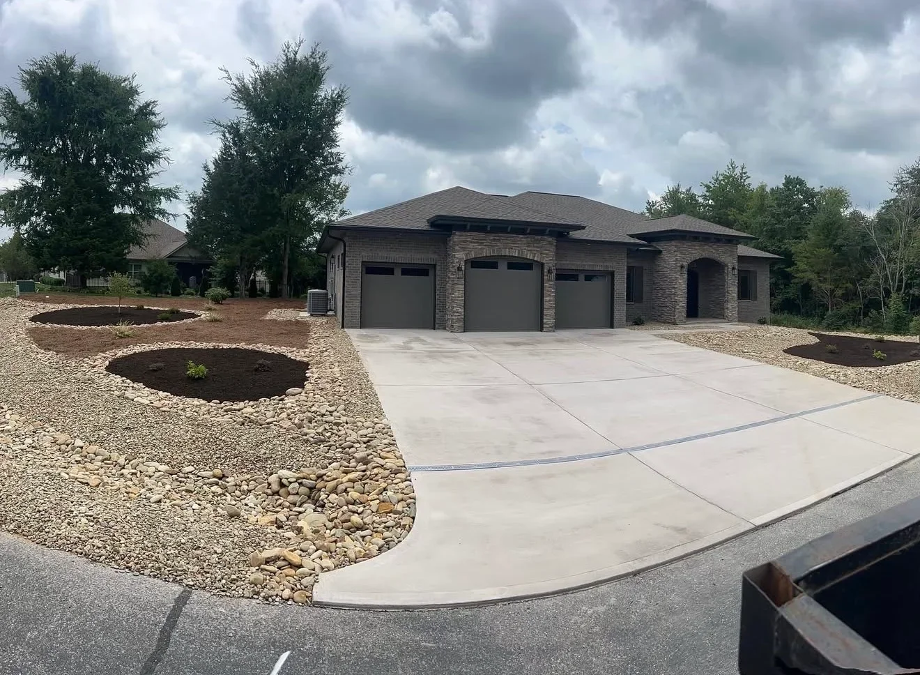 Concrete driveway with drainage channel, river rock borders, and mulched circular planting beds beside a brick ranch home