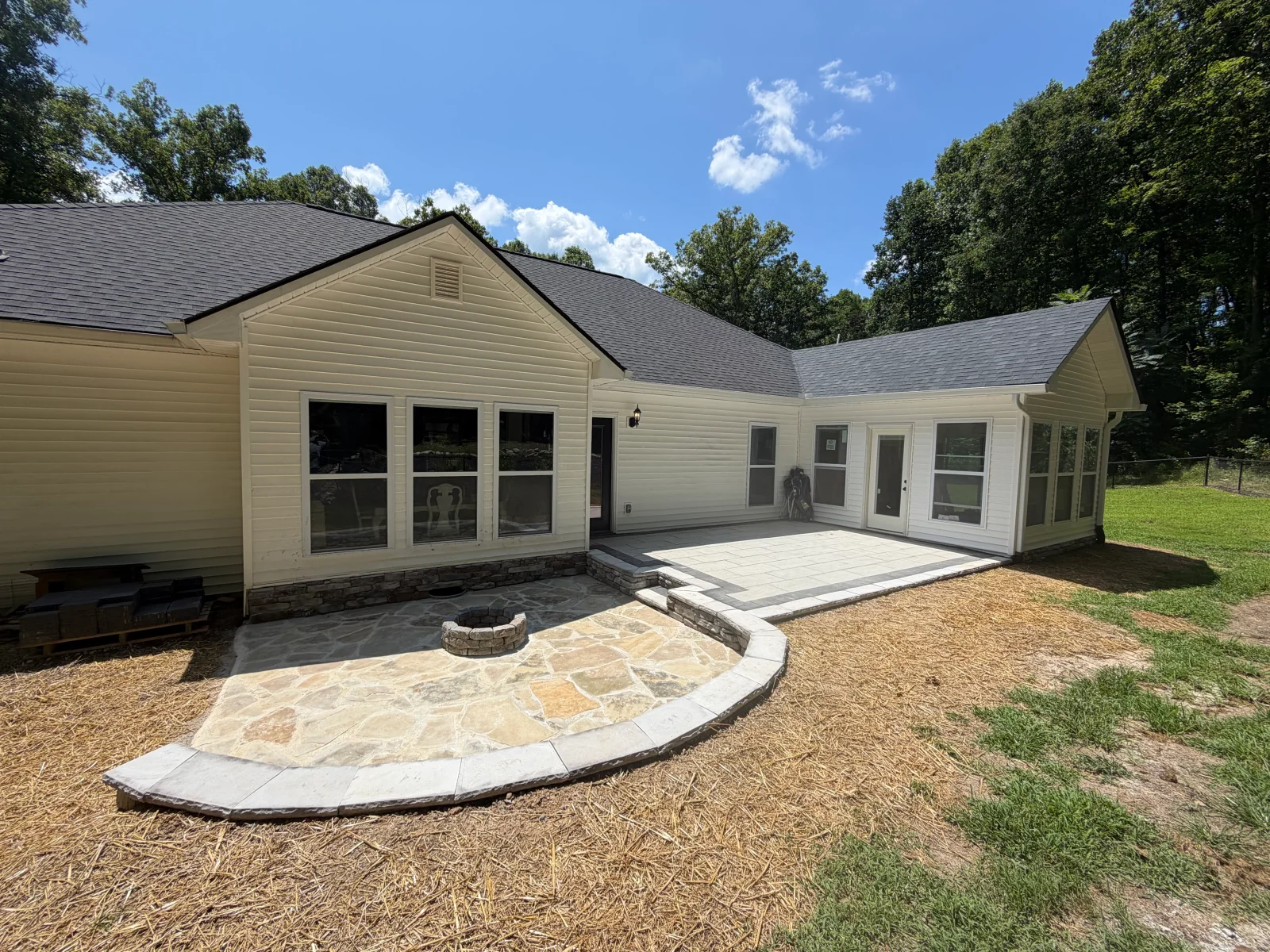 Flagstone patio with stone fire pit, curved paver edging, and gray paver patio beside a vinyl-sided house with stacked stone foundation