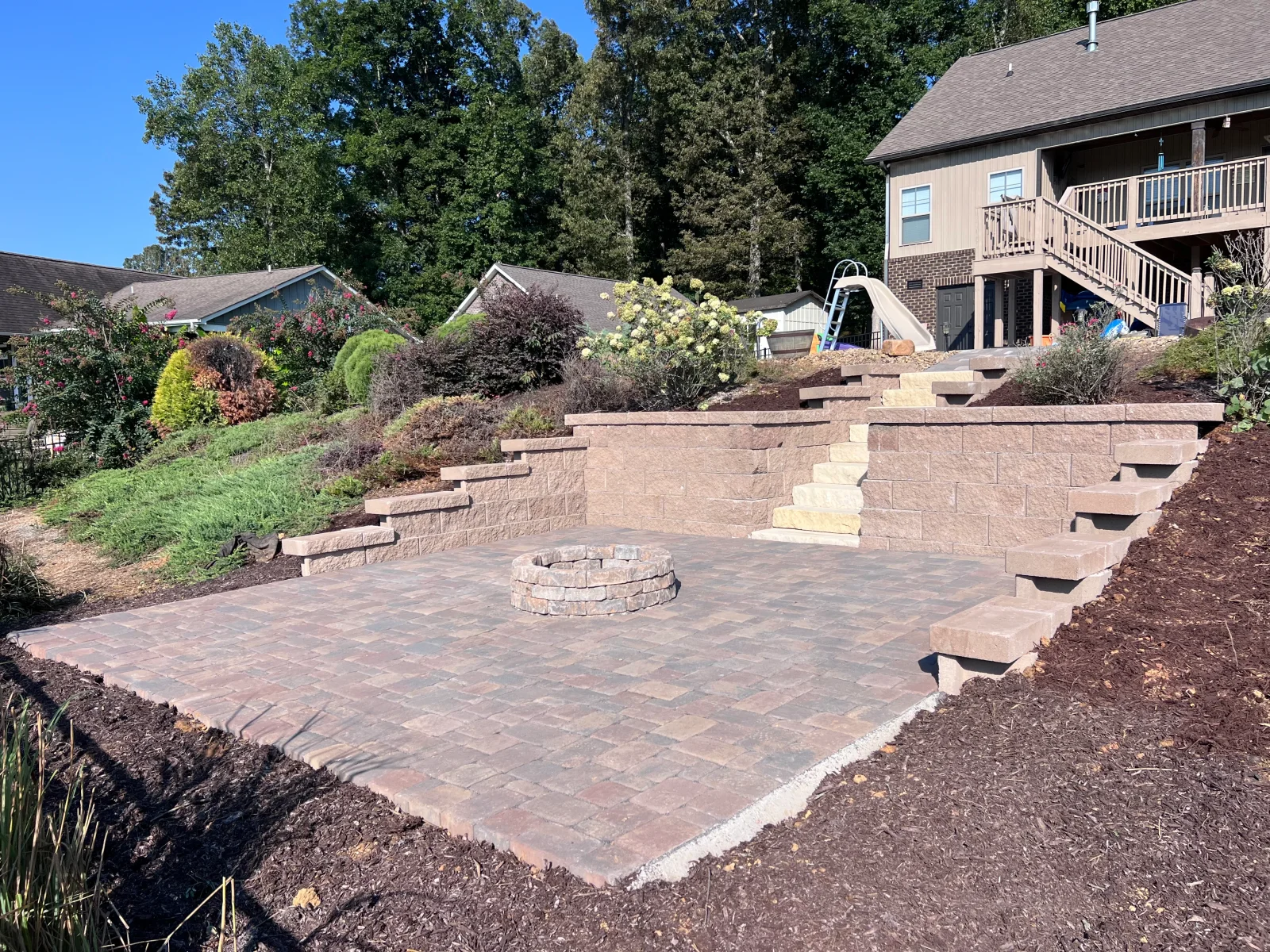 Tiered tan block retaining walls with paver patio, stacked stone fire pit, and block steps beside a sloped yard