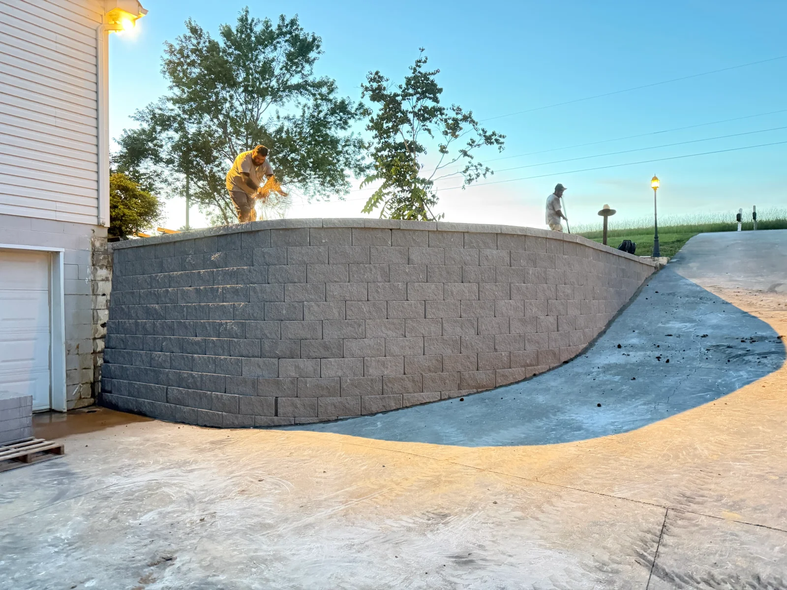 Curved gray split-face concrete block retaining wall beside a poured concrete driveway with workers placing top course