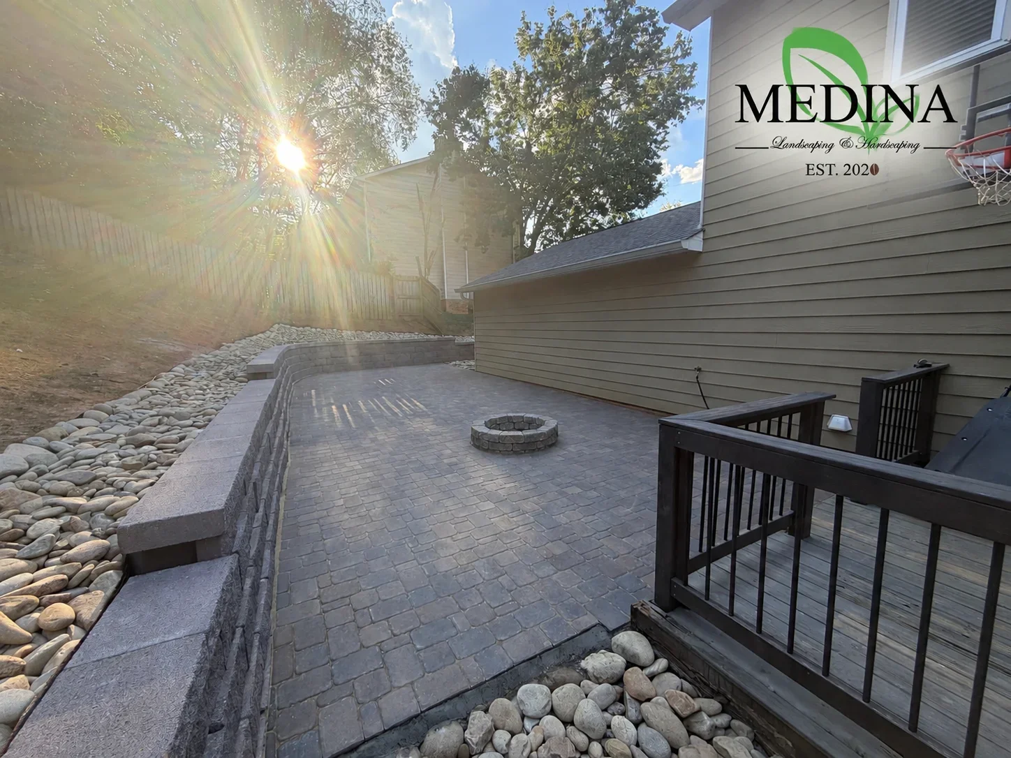 Gray interlocking paver patio with stacked-stone fire pit, segmental retaining wall, and river rock drainage bed beside a dark wood deck