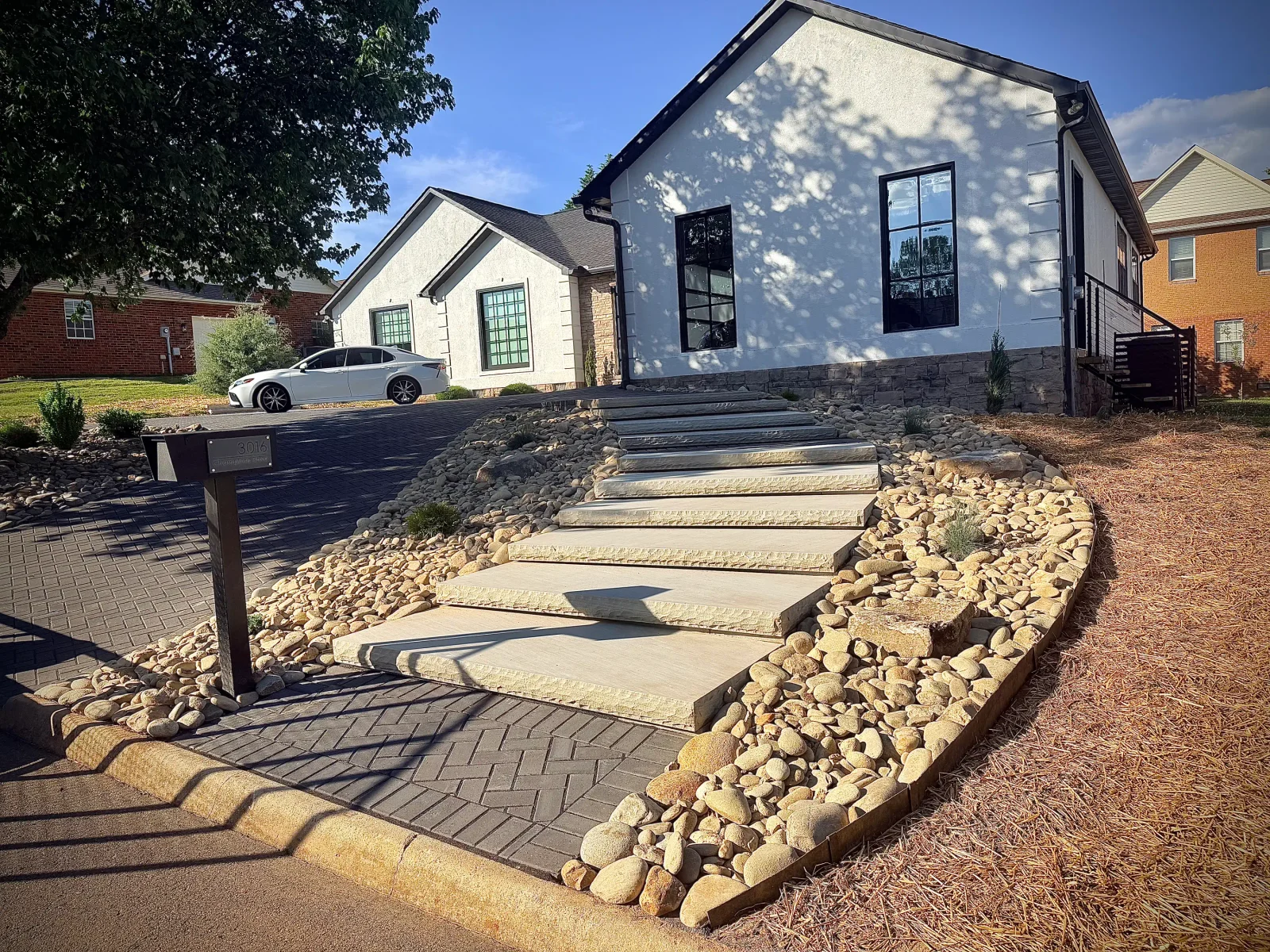Cut stone steps with river rock border, herringbone paver walkway, and black metal mailbox post in front yard